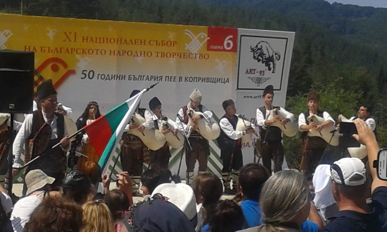 Гайдари в народни носии свирят на сцената на събора в Копривщица, до тях мъж развява българското знаме.