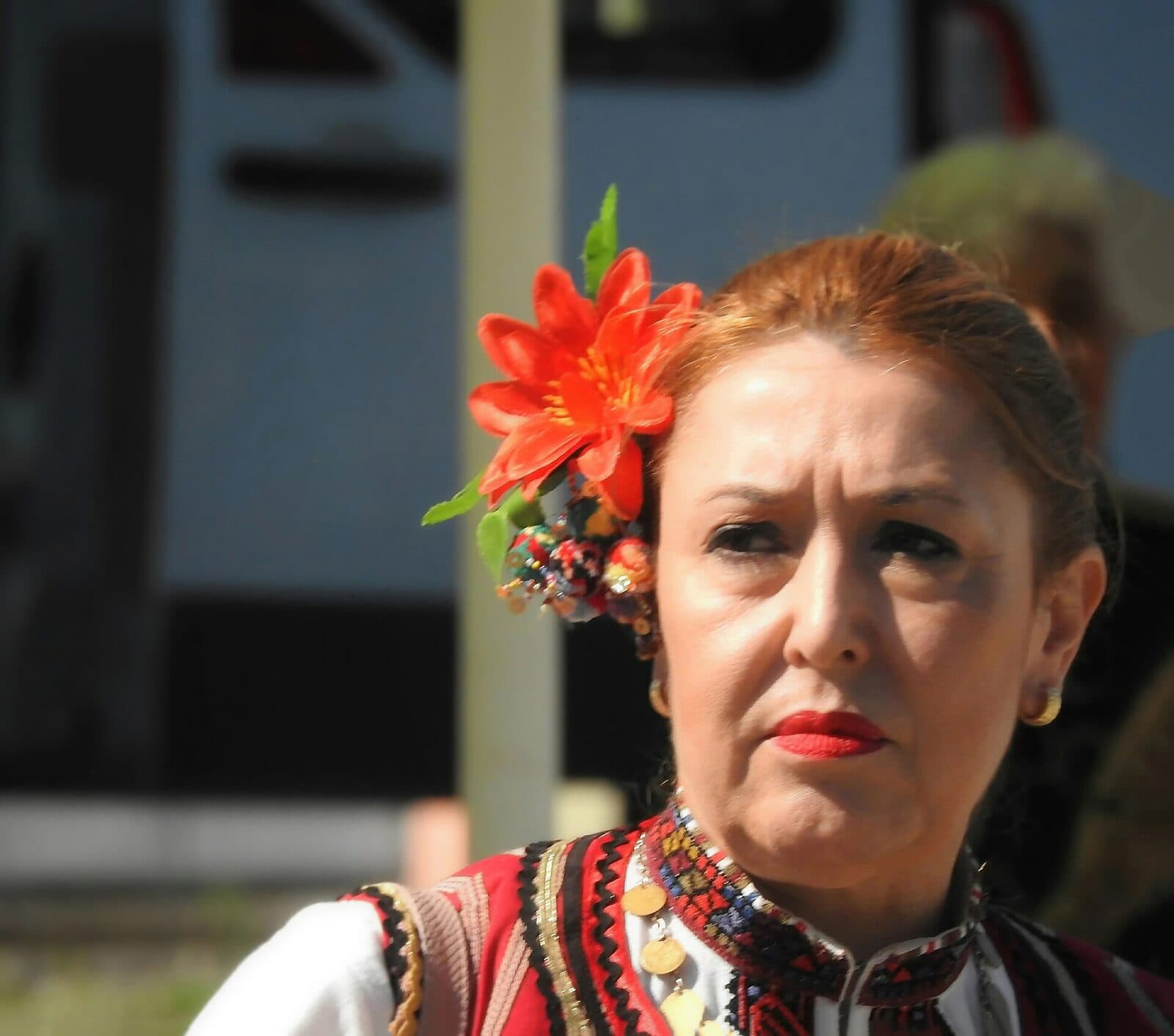 Жена в народна носия с червено цвете в косата, гледа встрани на фолклорен фестивал.
