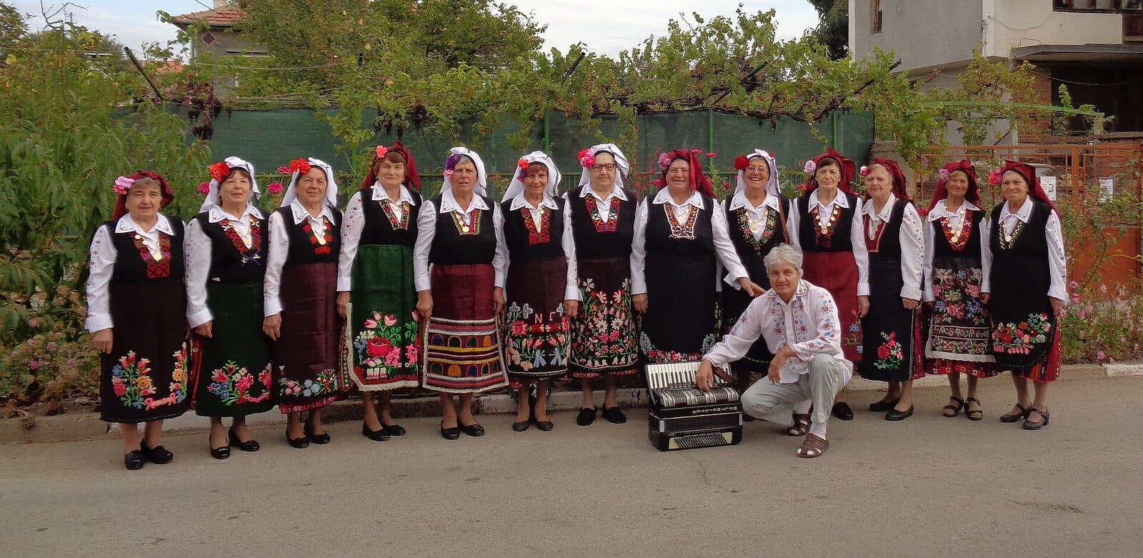 Жени в тракийски носии с черни сукмани и шарени престилки позират на открито, пред тях е приклекнал акордеонист.
