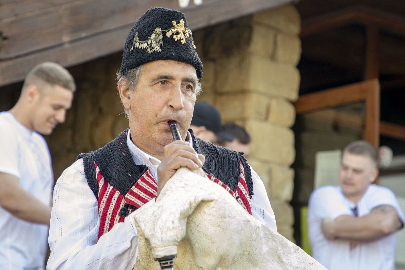 Възрастен гайдар в черен елек с червени ивици и калпак свири на гайда с бял козешки мях.