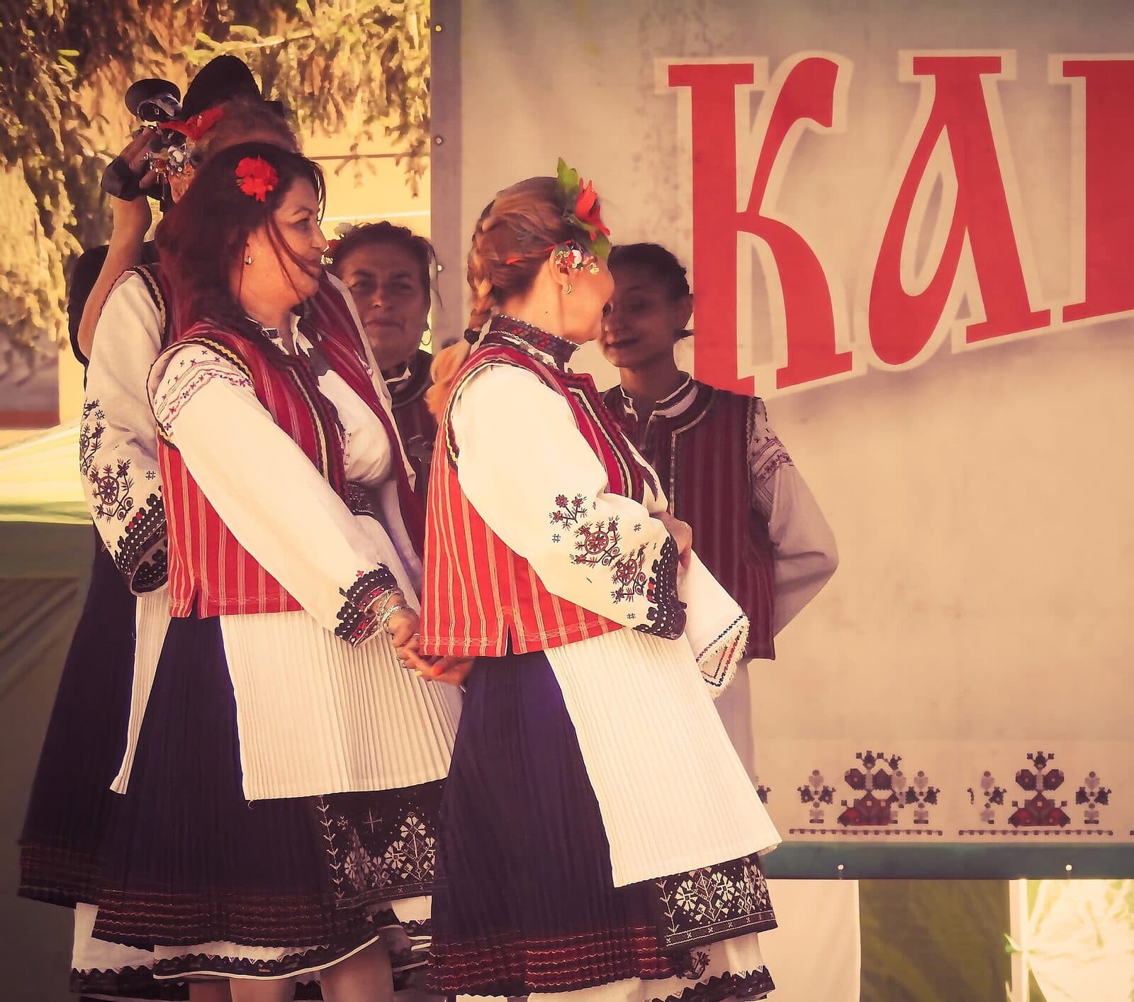 Две жени в капански носии с бели ризи, червени елеци и черни поли стоят на сцена на фолклорен празник.