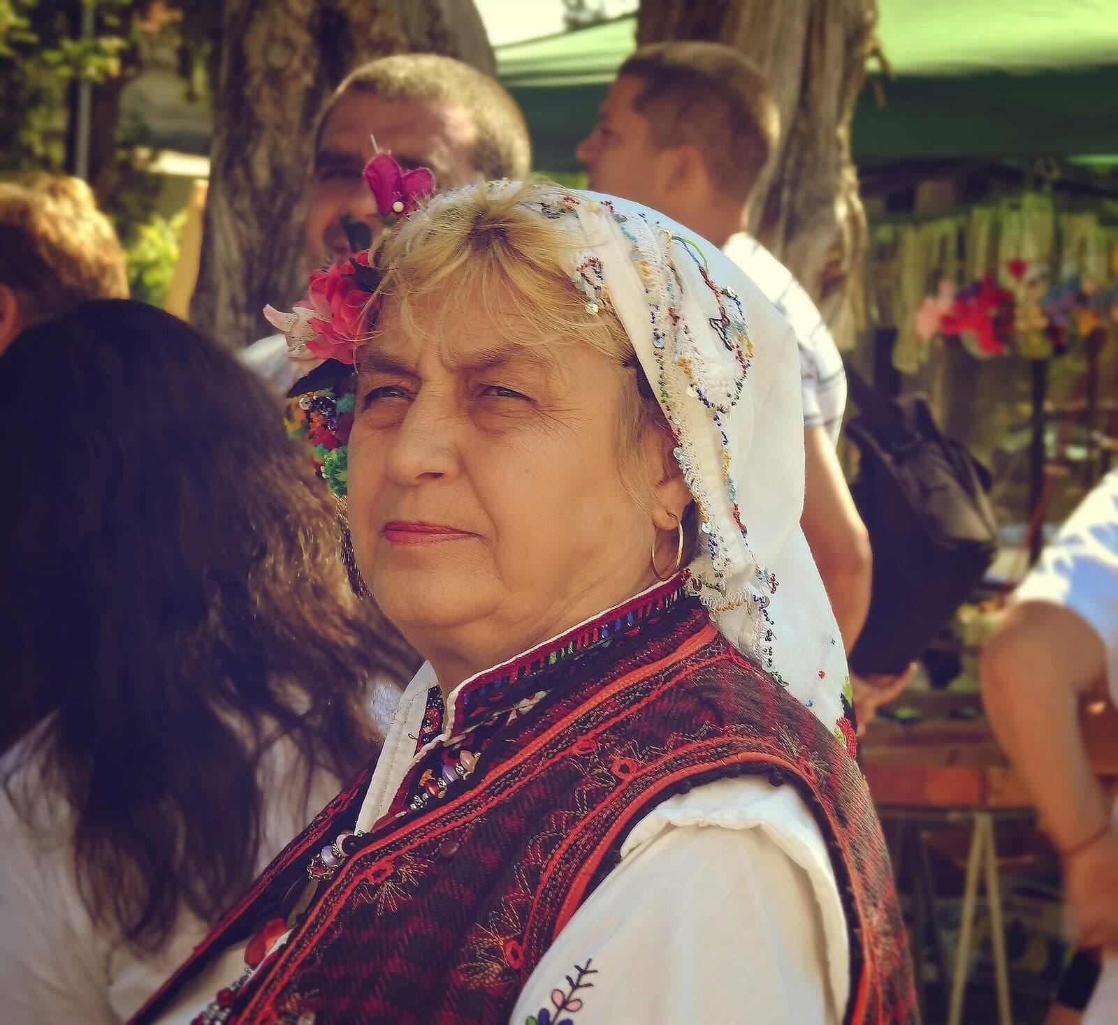 Възрастна жена в капанска носия с бяла забрадка и цветя гледа встрани на фолклорен празник.