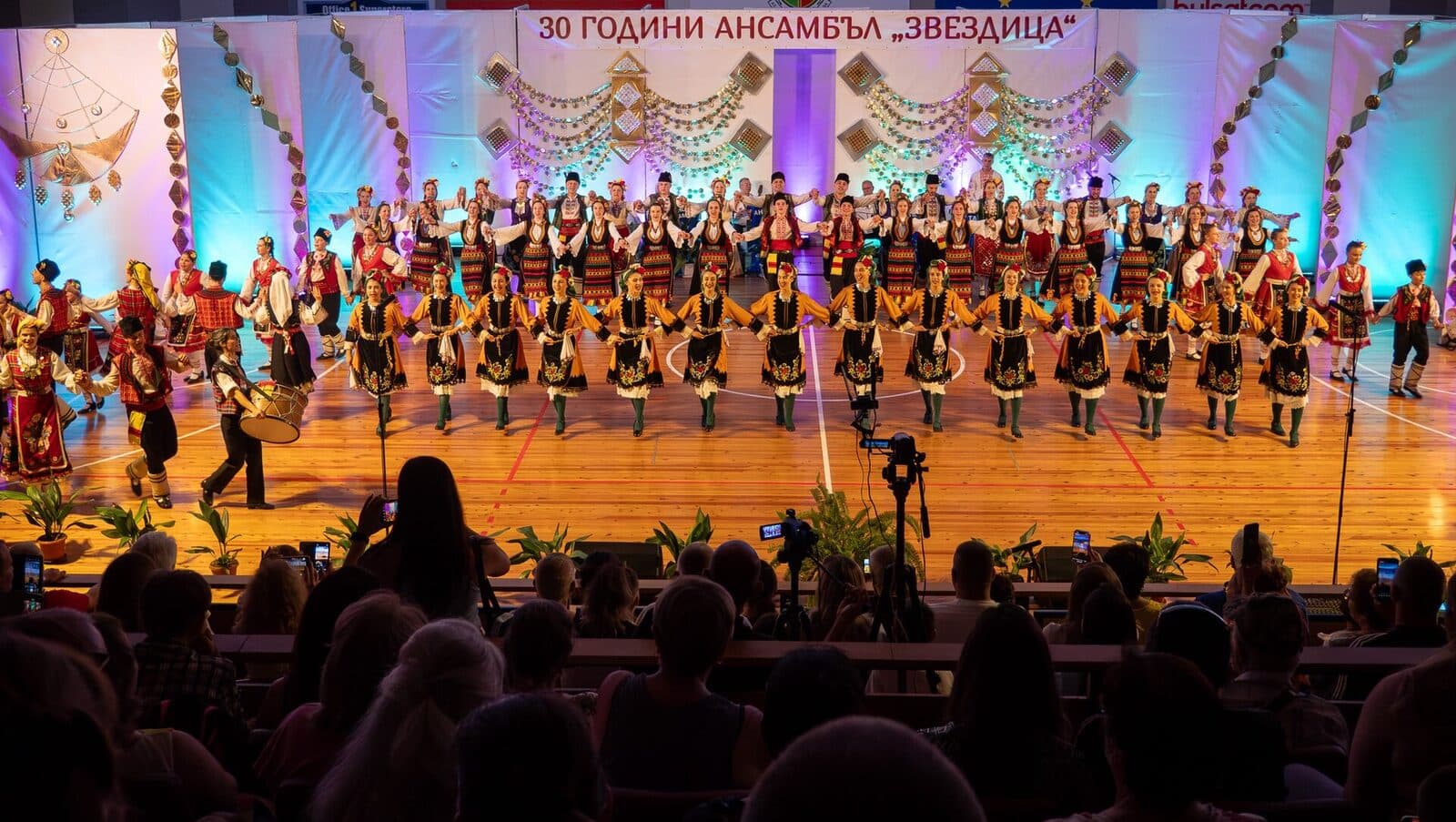 Голям танцов състав в тракийски носии играе хоро в няколко реда на сцена пред пълна зала.